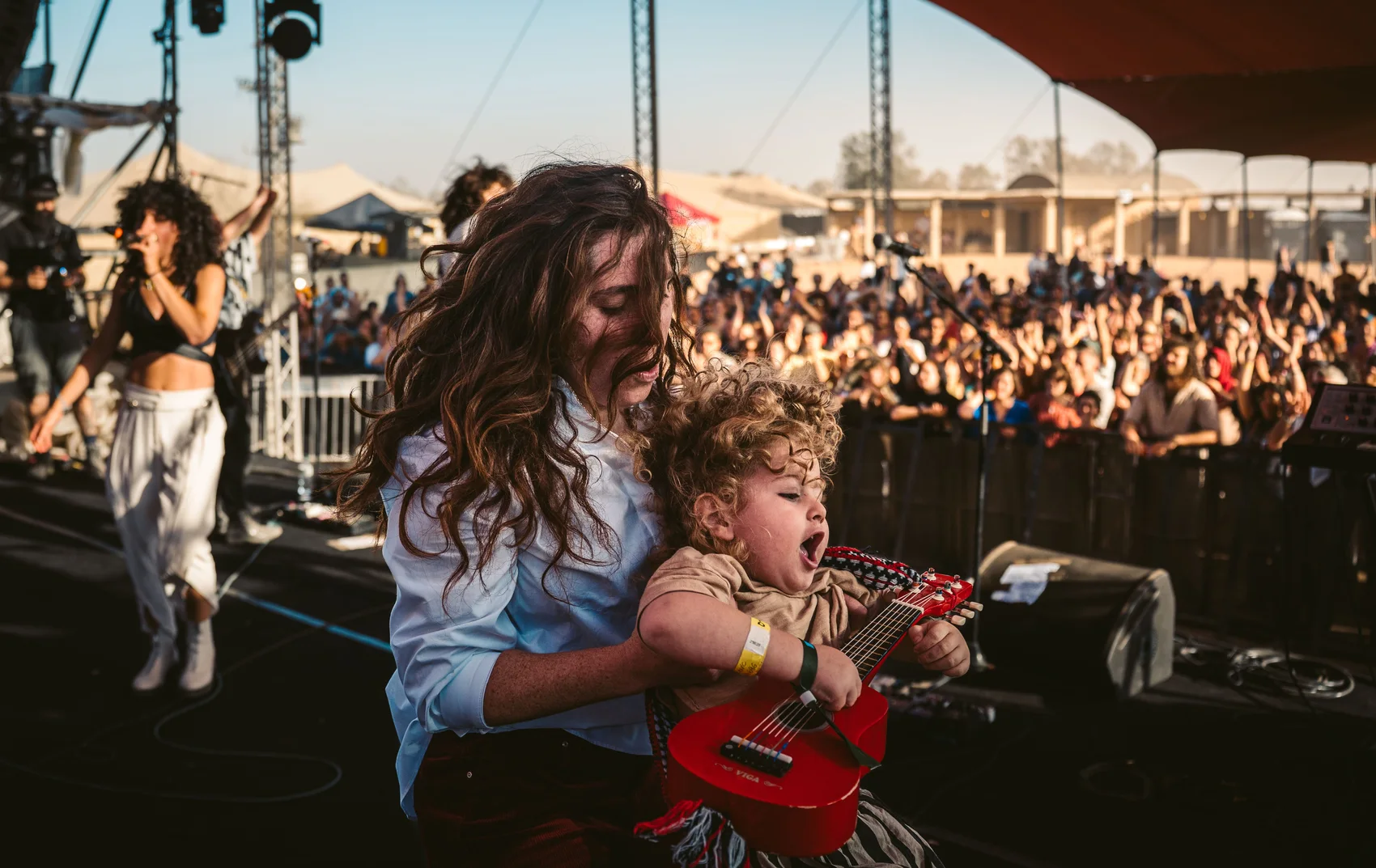 Hazeavot musician carrying a child and a small guitar beside the stage